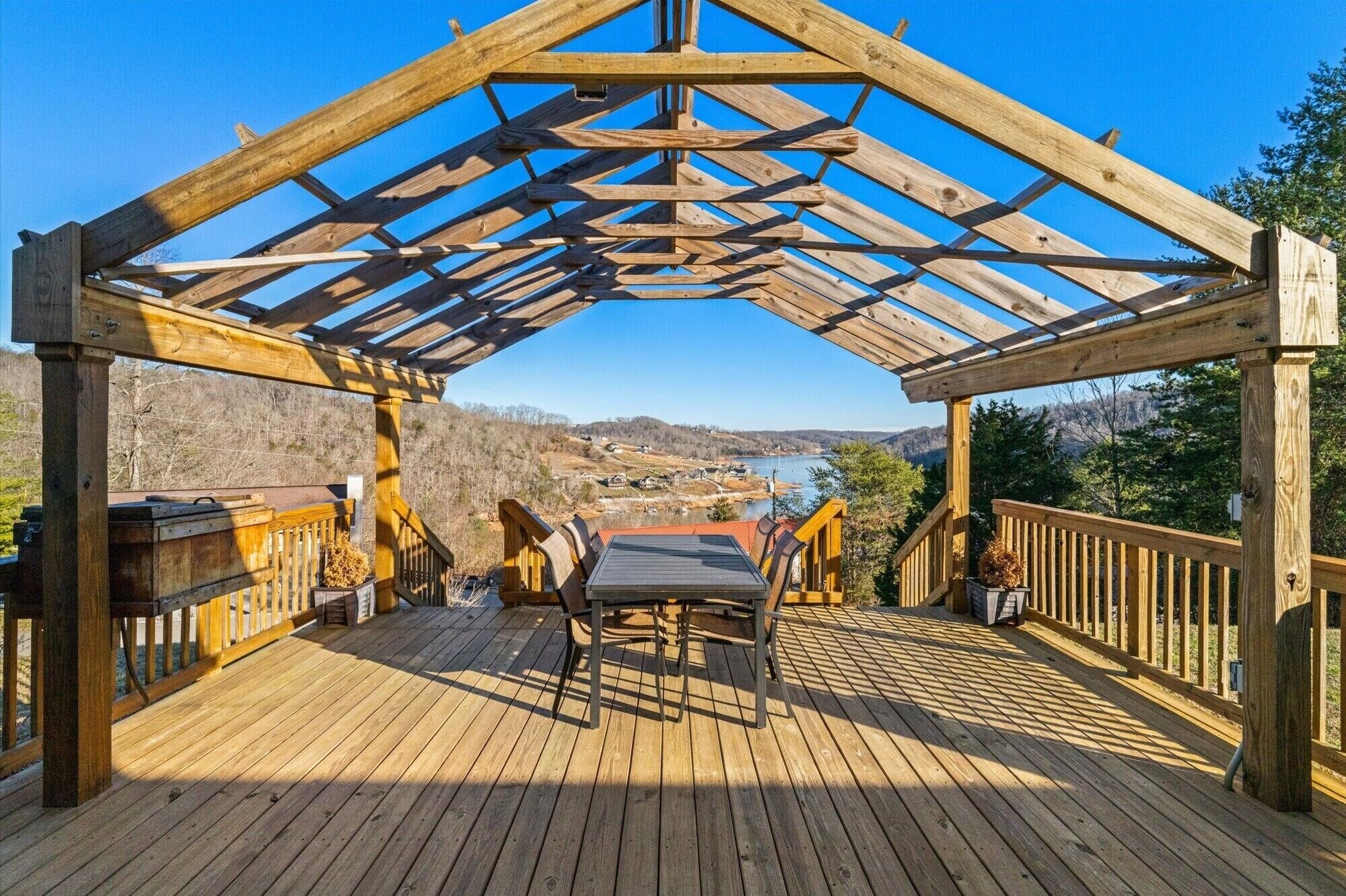 A wooden deck with a pergola structure, a table, and chairs, overlooking a scenic valley and lake.