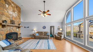 A spacious living room with a stone fireplace, leather sofas, a large window, and a ceiling fan.