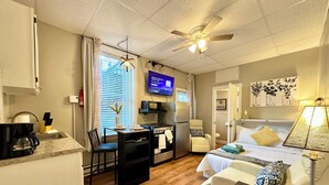 A compact hotel room with a kitchenette, a bed with two pillows, a desk with a lamp, and a ceiling fan.