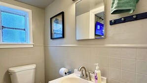 A bathroom with a toilet, sink, and mirror. There is a towel rack with a green towel, a framed picture on the wall, and a window with a view of the outside.