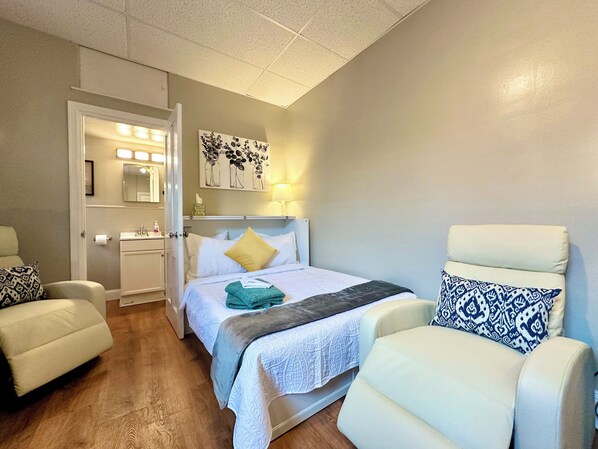 Habitación de hotel con cama, dos sillones, una mesita de noche, un espejo y una obra de arte en la pared.