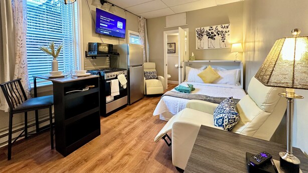 A hotel room with a bed, a desk with a computer, a microwave, a refrigerator, a ceiling fan, and a TV displaying a welcome message.