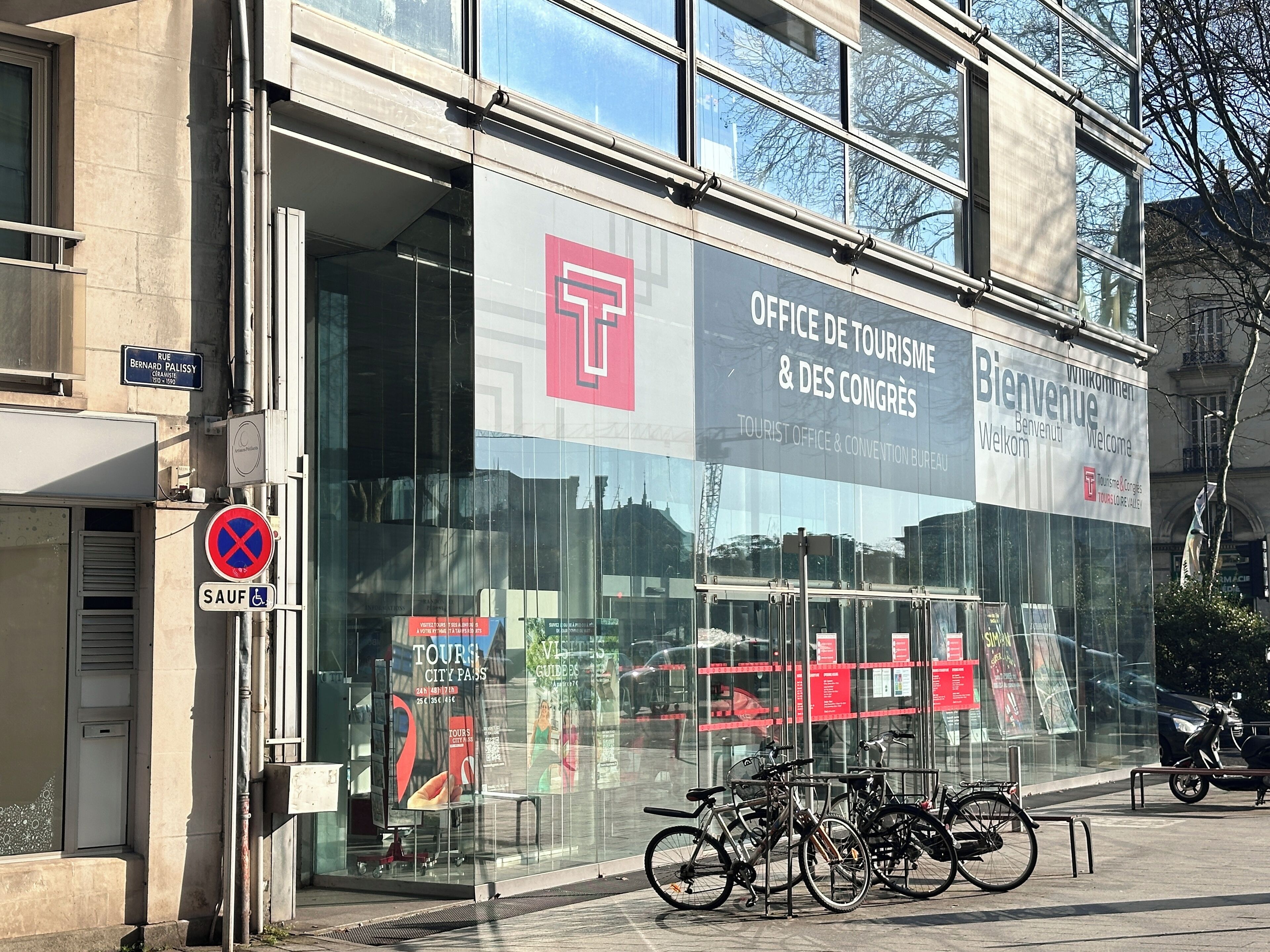 Um edifício moderno com fachada de vidro extensa, sinalização do Office de Tourisme & des Congrès e bicicletas estacionadas na frente.