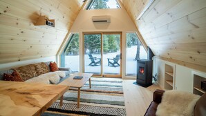 Ein gemütlicher Raum mit Holzwänden, einer schrägen Decke, einem Kamin und einem großen Fenster mit Blick auf eine verschneite Landschaft.