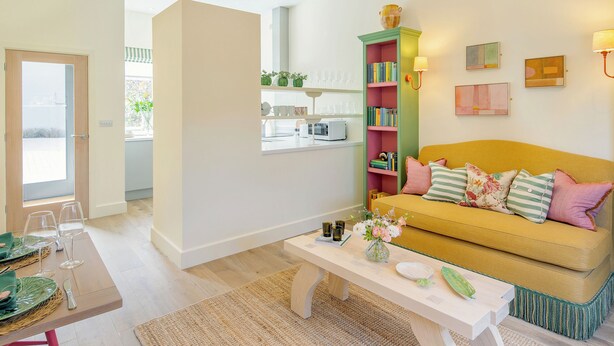 Una sala de estar con un sofá amarillo, una mesa de comedor con sillas, una estantería y una ventana con vistas.