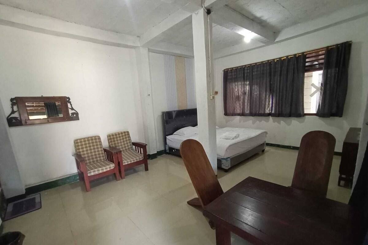 A room with a bed, two chairs, a table, a window with curtains, and a wall-mounted shelf.