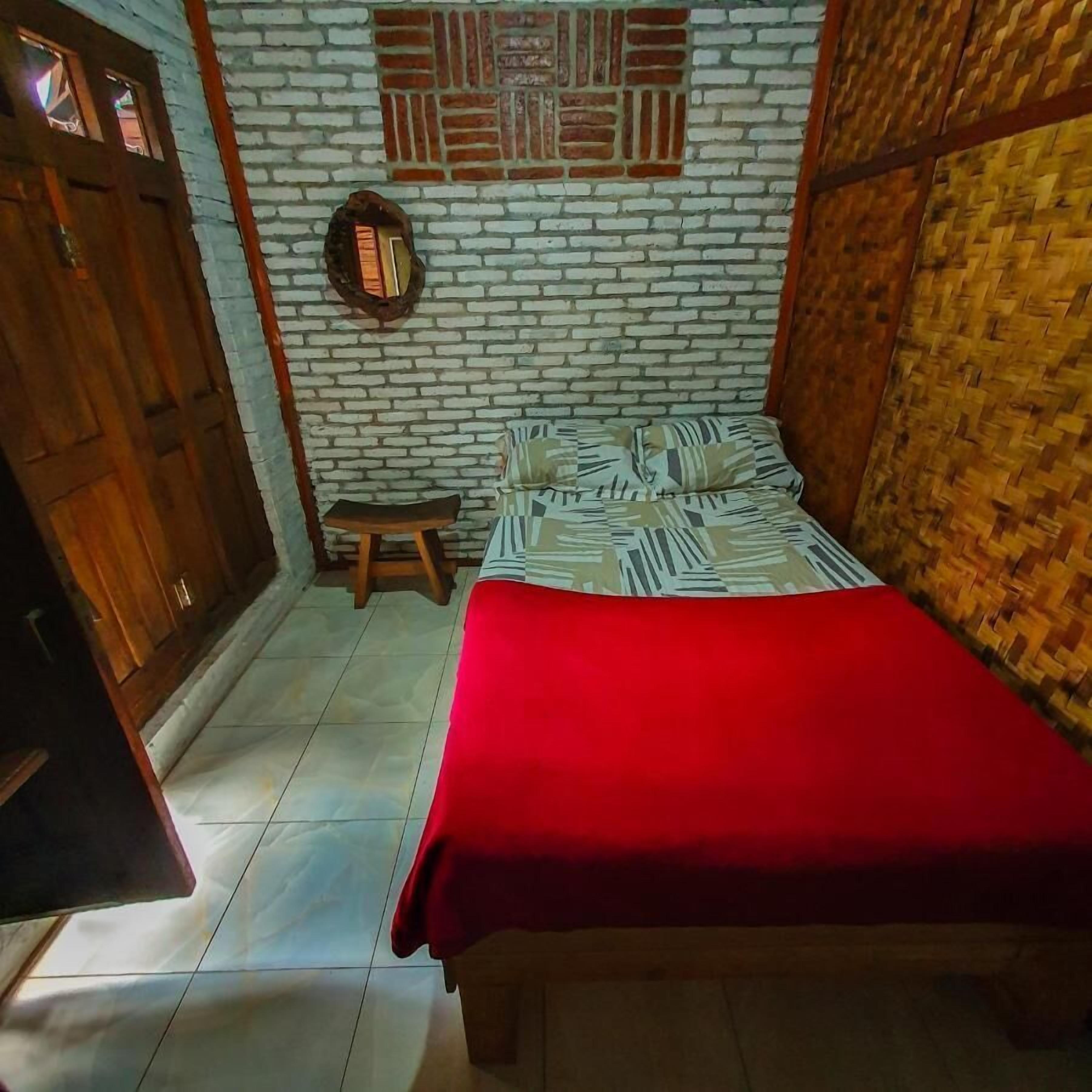 A room with a bed, a small stool, a wooden door, and a brick wall with a decorative panel.