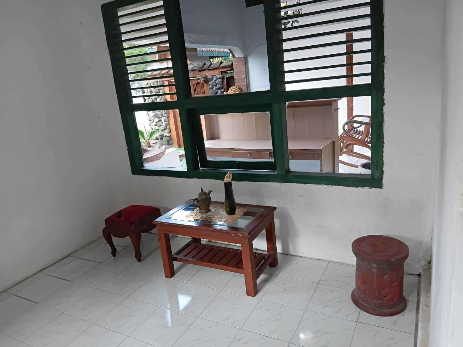 A room with a tiled floor, a small wooden table with a teapot, a red stool, and a window with green shutters.