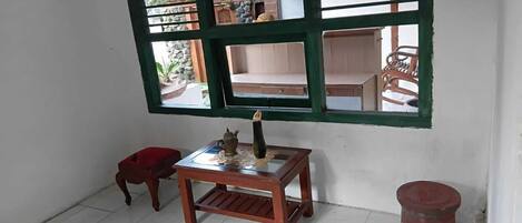 A room with a tiled floor, a small wooden table with a teapot, a red stool, and a window with green shutters.