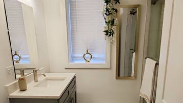A bathroom with a toilet, sink, vanity, mirror, window with blinds, and a small wooden mat on the floor.