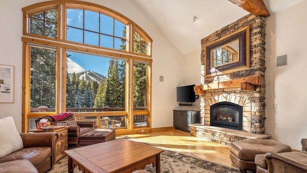 A cozy living room with a stone fireplace, large windows, and a view of a snowy mountain.