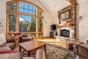 Una sala de estar acogedora con una chimenea de piedra, ventanas amplias y vistas a una montaña nevada.
