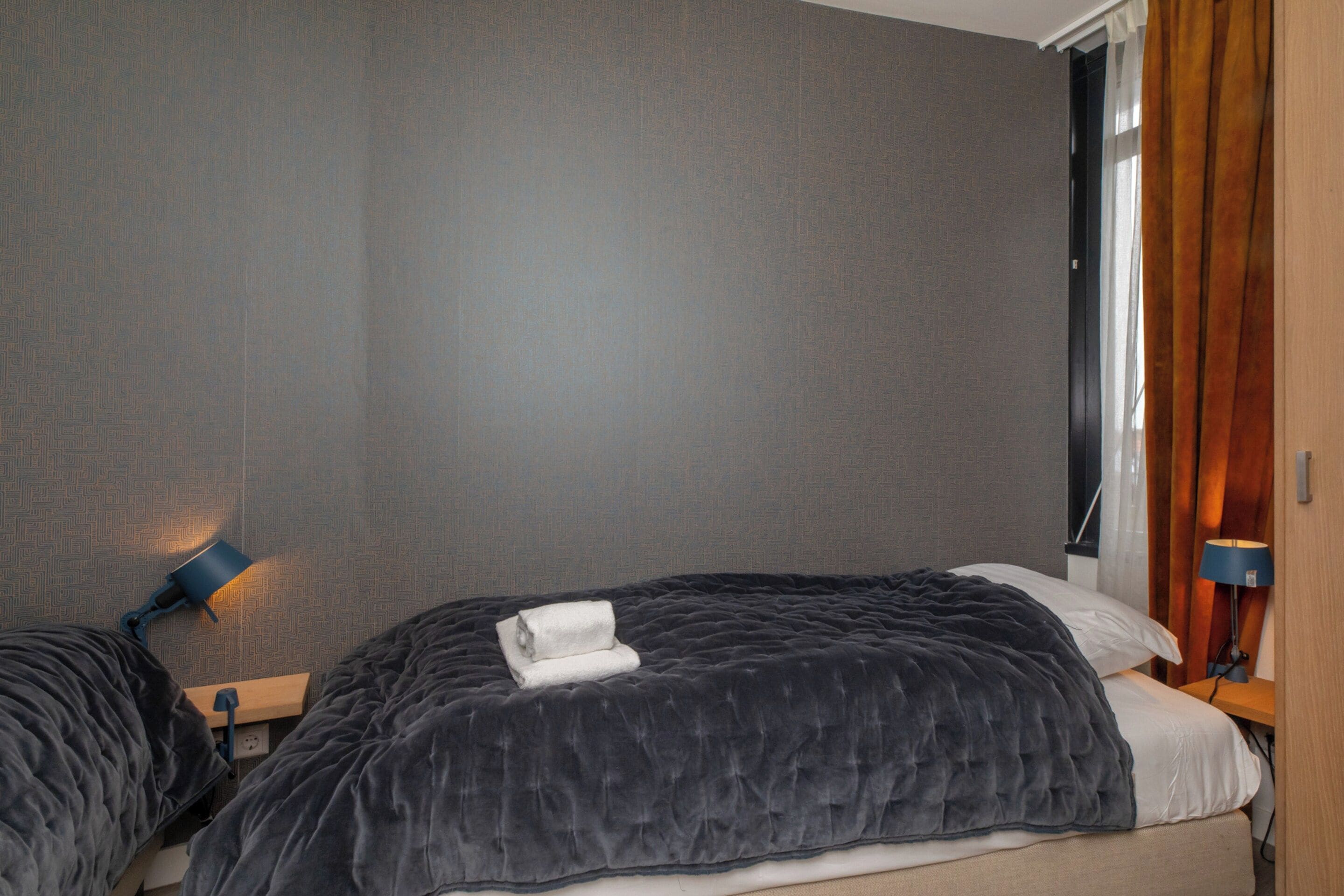 Una camera d'hotel con un letto, una lampada da comodino, una finestra con tende e una parete con motivo geometrico.