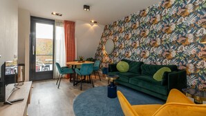 A modern living room with a green sofa, yellow armchair, and blue round rug. A dining area with teal chairs and a table is visible. The room features a colorful floral wallpaper and a large mirror on the wall. - Wifi and Golden Beaches; Home (Zoutelande)