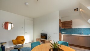 A modern living room with a round wooden dining table, two blue chairs, a yellow armchair, and a small TV on a wooden cabinet. - Wifi and Golden Beaches; Home (Zoutelande)