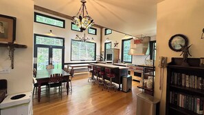 A spacious kitchen with a large island, wooden flooring, and a chandelier. There are multiple windows providing natural light and views of the outdoors.