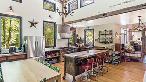 A spacious kitchen with large windows, a central island with stools, and a dining area with a wooden table.