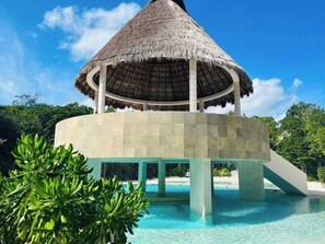 Estructura de techo de paja con piscina debajo, rodeada de follaje y un cielo despejado.
