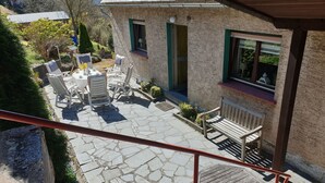 Um pátio de pedra com mobiliário de exterior, um banco de madeira e uma casa com janelas emolduradas a verde.