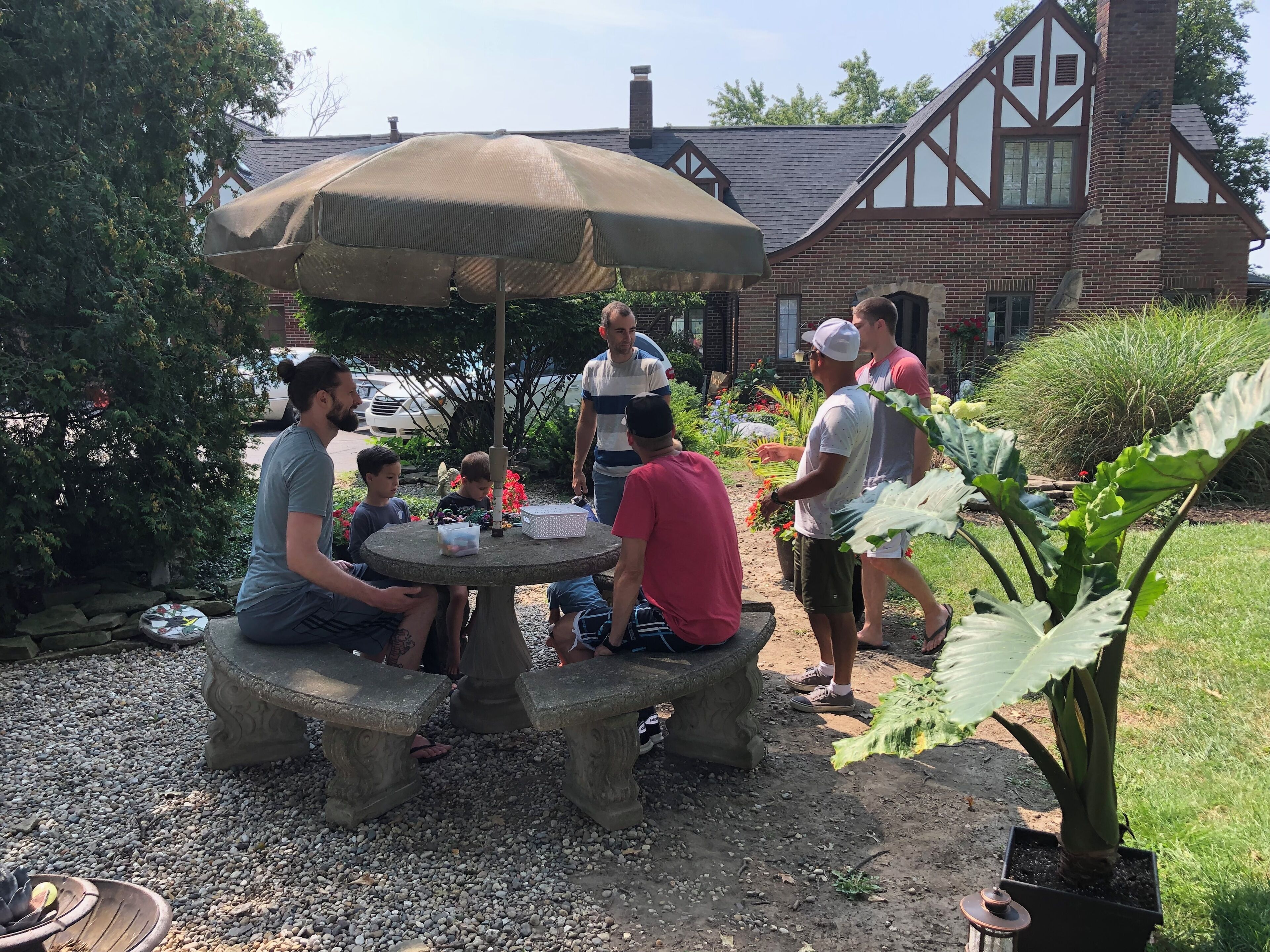 Un groupe de personnes est assis autour d’une table en pierre, protégée par un parasol, dans un jardin où l’on aperçoit une maison au fond.