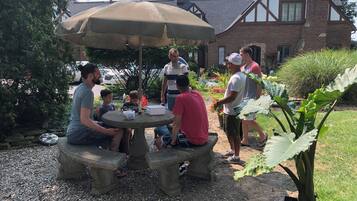 Un groupe de personnes est assis autour d’une table en pierre, protégée par un parasol, dans un jardin où l’on aperçoit une maison au fond.