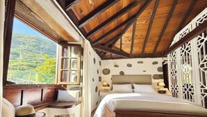 A bedroom with a wooden ceiling, a bed, a bench with cushions, and a view of greenery outside.