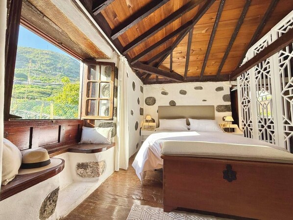 A bedroom with a wooden ceiling, a bed, a bench with cushions, and a view of greenery outside.