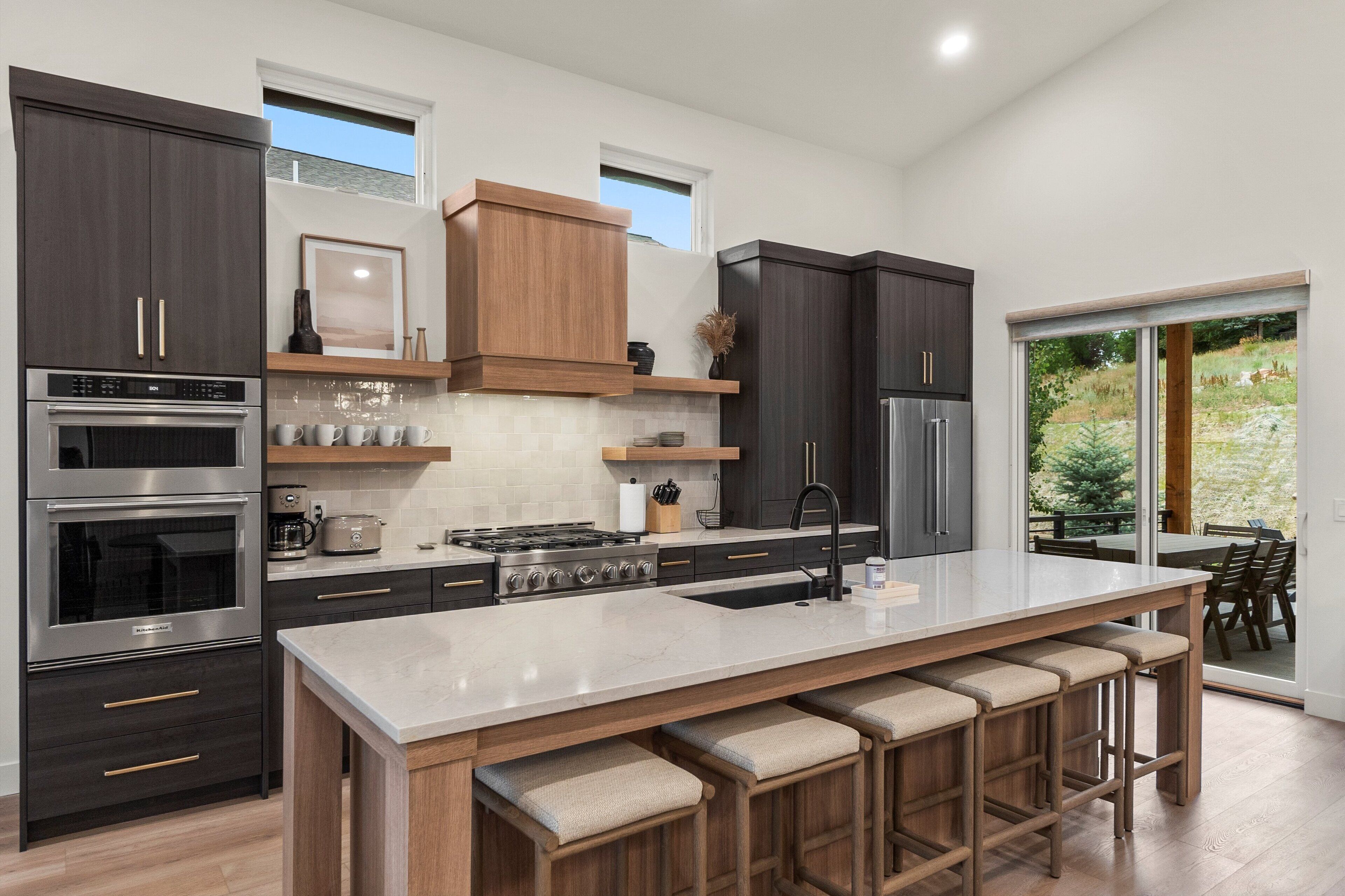 A modern kitchen with dark cabinetry, a large island with bar stools, and a view of the outdoors through a sliding glass door.