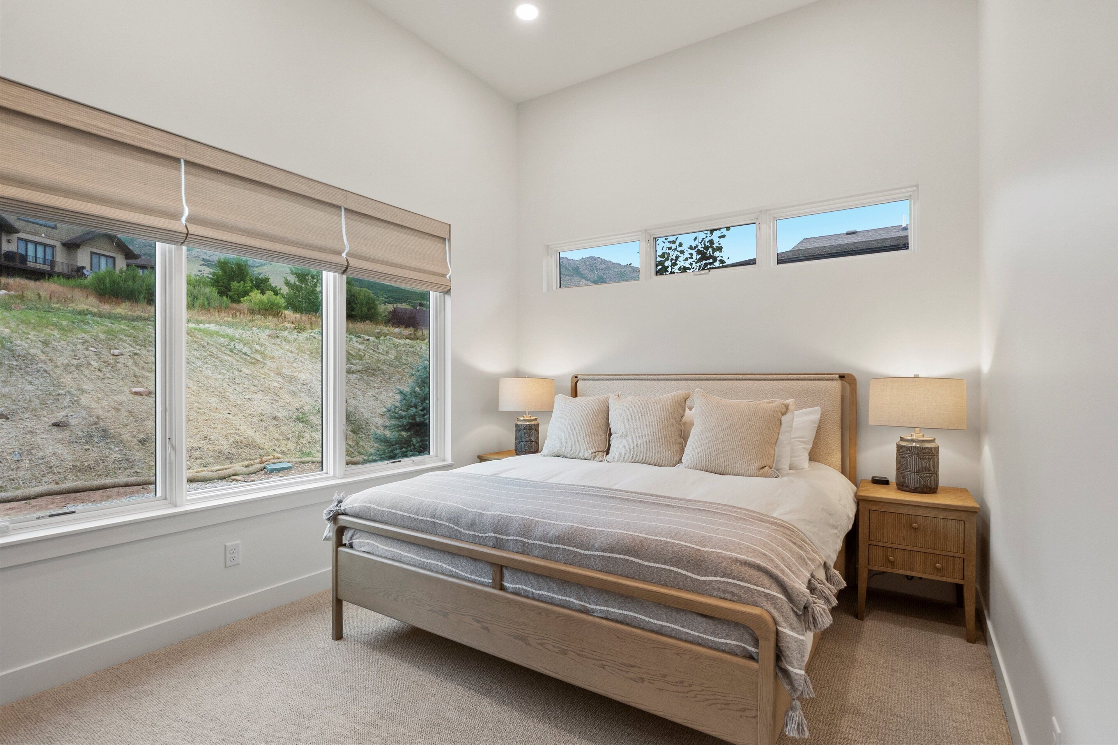 A bedroom with a bed, two bedside tables with lamps, a window with blinds, and a view of a landscape.