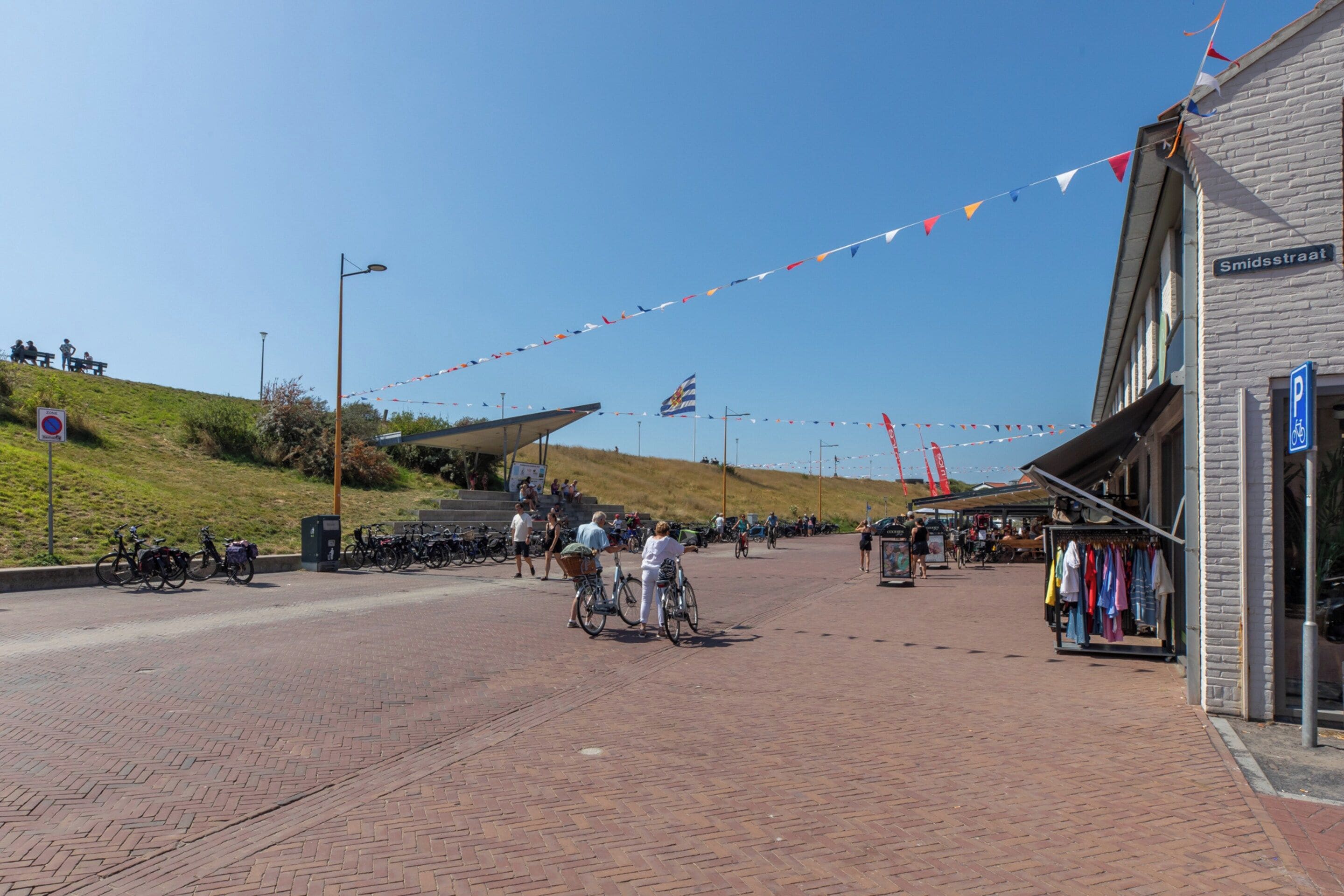 A paved pedestrian area with a row of shops on one side and a grassy hill on the other.
