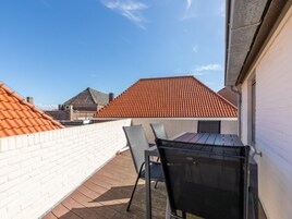 A rooftop terrace with a table and chairs, a white brick wall, and red-tiled roofs in the background.