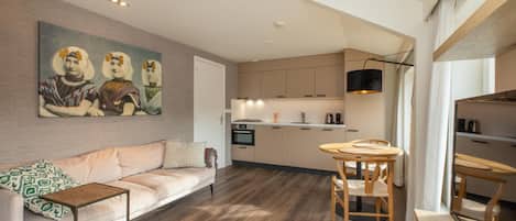 A modern living room with a sofa, a coffee table, and a dining area with a table and chairs. There is a kitchen in the background with cabinets and a stove.