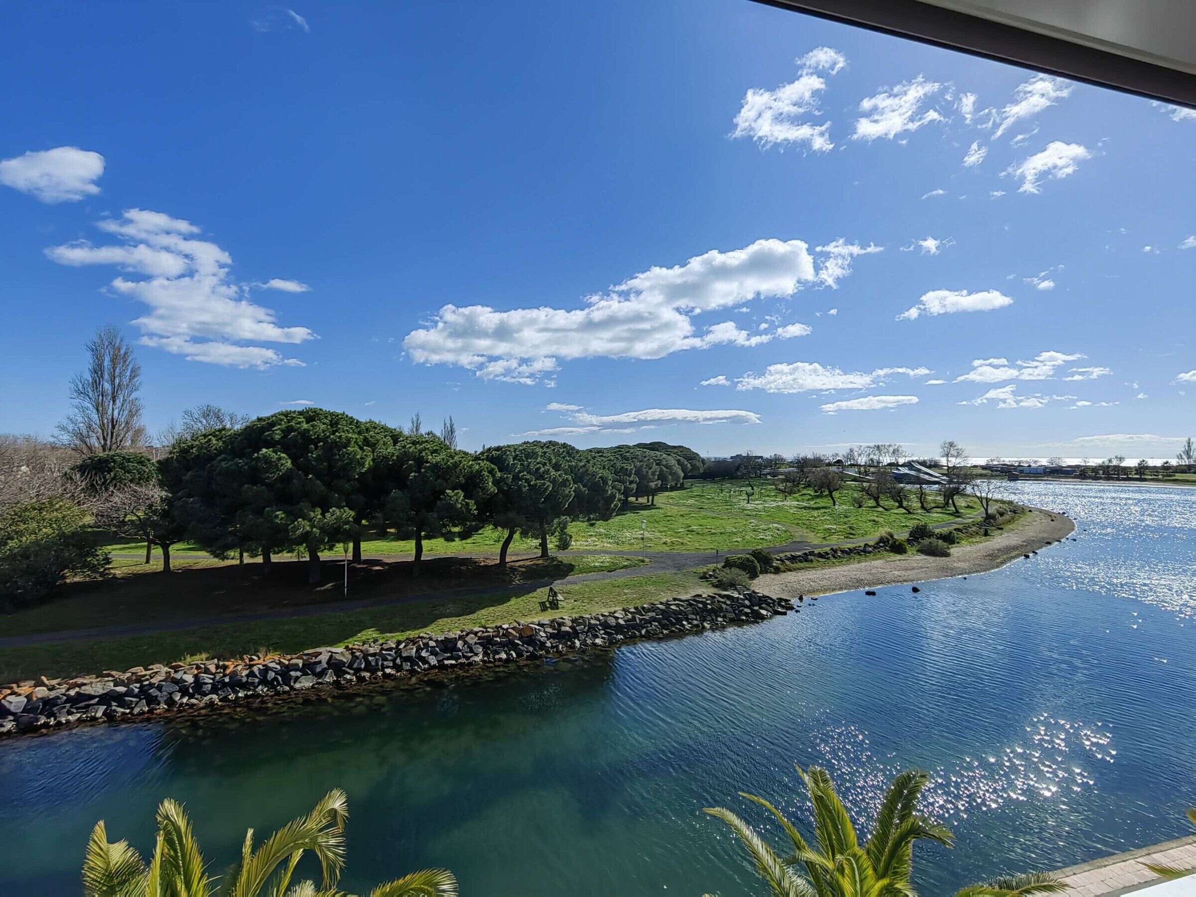 A river with a stone wall and trees on the bank, a clear sky, and a distant view of buildings.