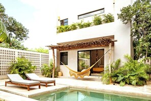 Una casa moderna con piscina, dos sillones de descanso, una hamaca y abundante vegetación.