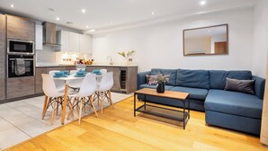 Eine moderne Küche mit weißen Schränken, ein Essbereich mit Tisch und Stühlen sowie ein blaues Sofa im Wohnzimmer.
