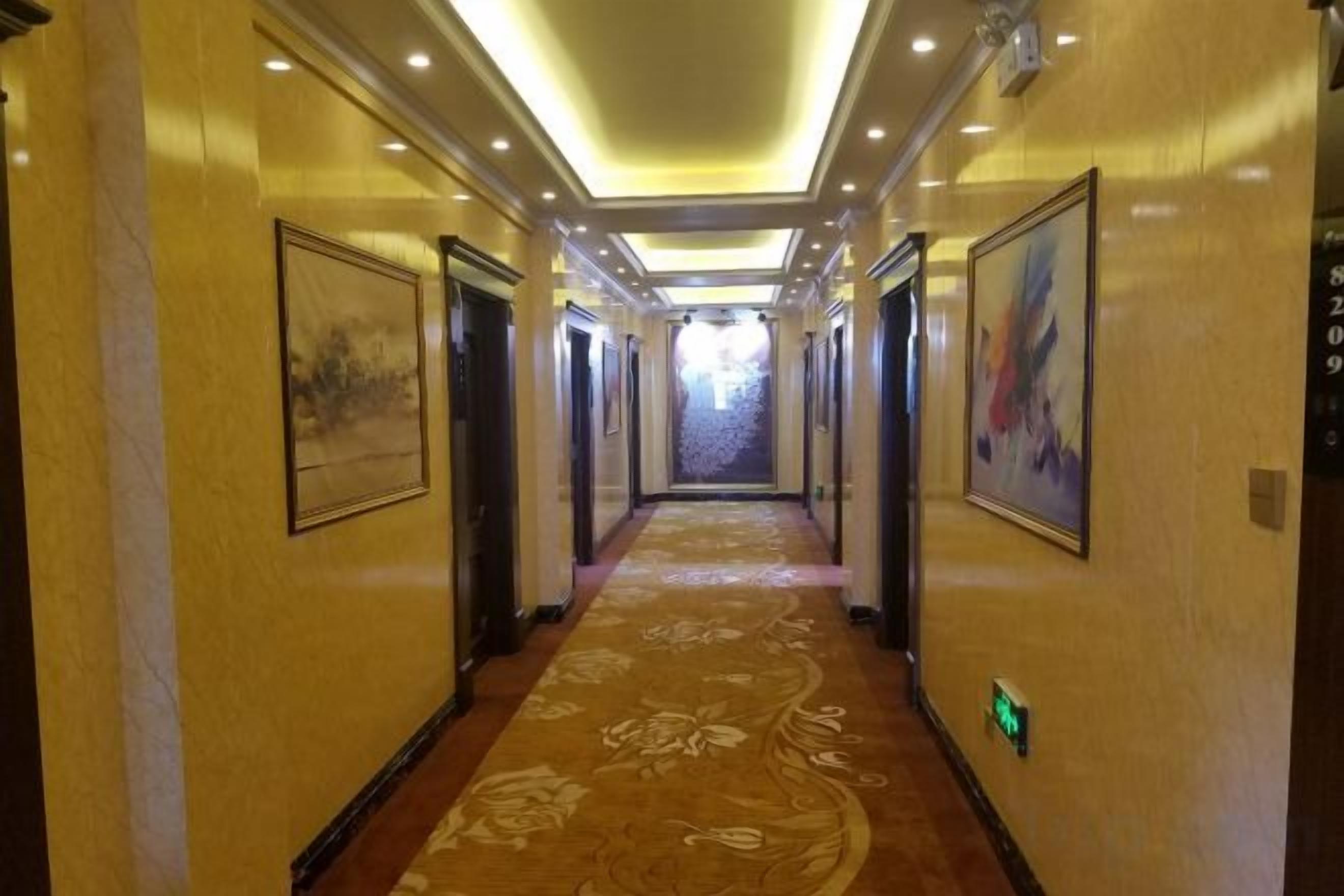 A hotel corridor with framed artwork on the walls, a carpeted floor, and a green exit sign.