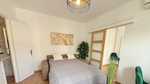 A bedroom with a bed, a bedside table with a plant, a wall-mounted artwork, and a door leading to another room. - 06CP - Beautiful T2 super equipped with terrace (Villeneuve-Loubet)