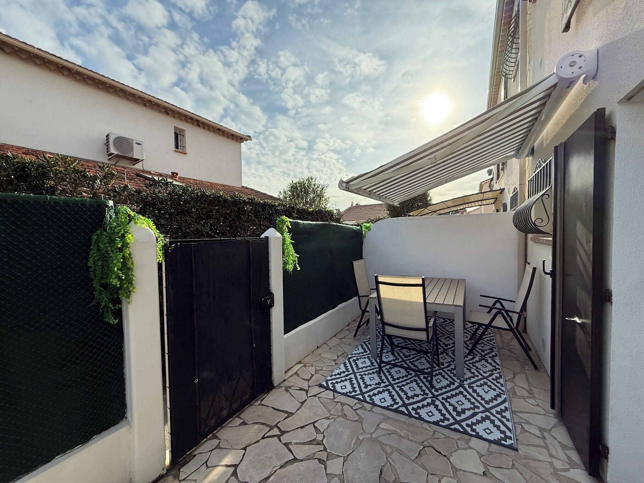 Une terrasse avec une table et des chaises, un tapis à motifs, un portail noir et une clôture verte.