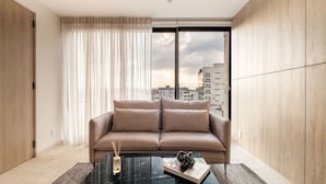 Un salon moderne avec un canapé beige, une table basse noire sur laquelle se trouvent un livre et un objet décoratif, et une vue sur la ville grâce à de grandes fenêtres.