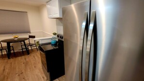 Una cocina con un refrigerador de acero inoxidable, una mesita con sillas y un ventanal con persianas.
