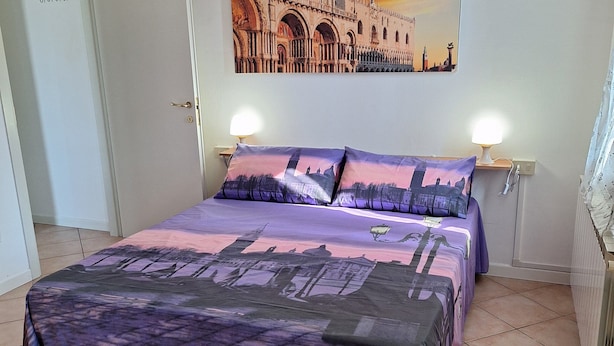 Una cama bien hecha con una colcha de diseño urbano en tonos morado y negro, dos lámparas de noche y un cuadro de gran formato con un paisaje urbano en la pared.