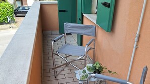 Un pequeño balcón exterior con dos sillas de metal y una mesa, una planta en maceta y una puerta verde al fondo.