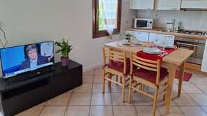 Cocina moderna con armarios blancos, microondas y estufa a gas. Hay una mesa de comedor con sillas y un televisor mostrando un noticiero.