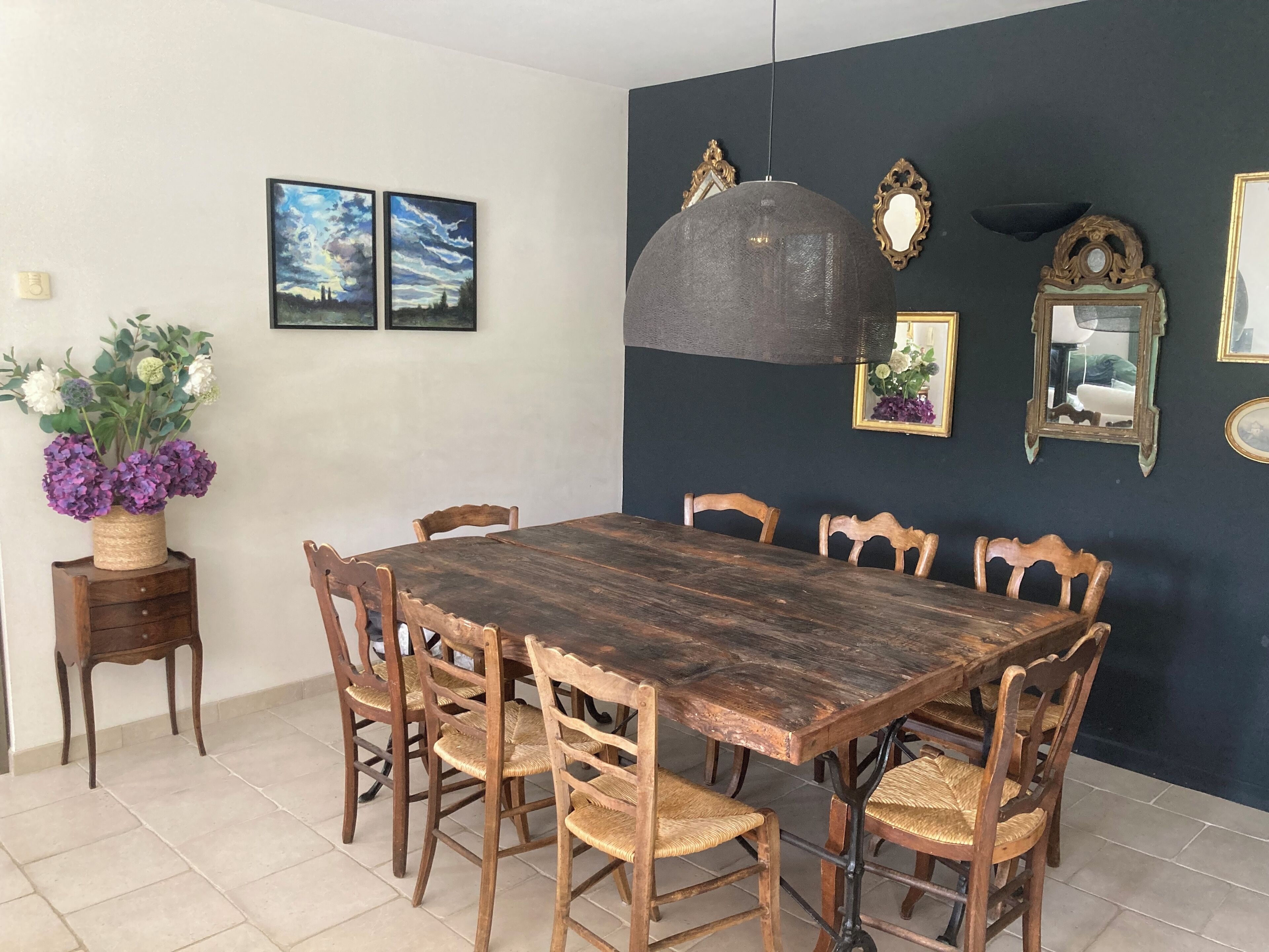 Un comedor con mesa y sillas de madera, una lámpara colgante y espejos y cuadros en la pared.