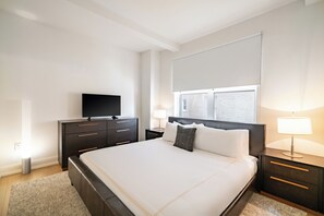 Una habitación de hotel moderna con una cama grande, un televisor de pantalla plana, una mesita de noche con lámpara y una ventana con persianas.