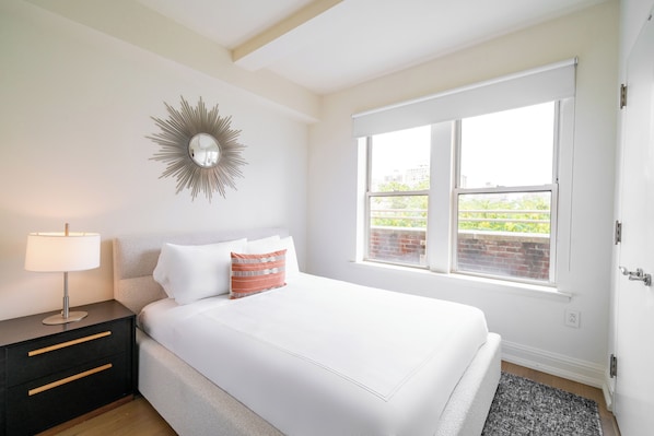 A bedroom with a bed, a nightstand, a lamp, a window, and a wall decoration.