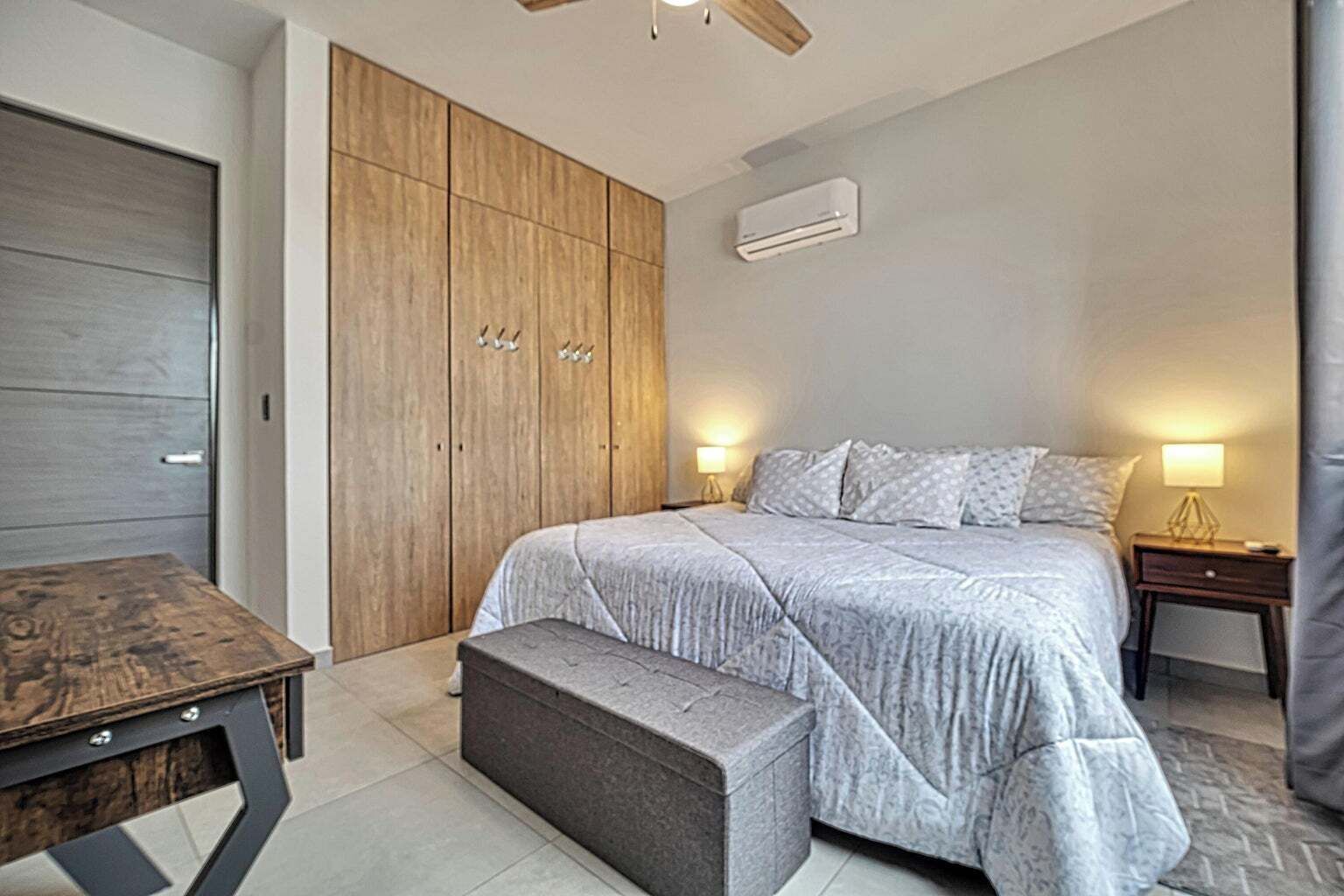 A bedroom with a bed, bedside tables, a bench, a wardrobe, and an air conditioning unit.