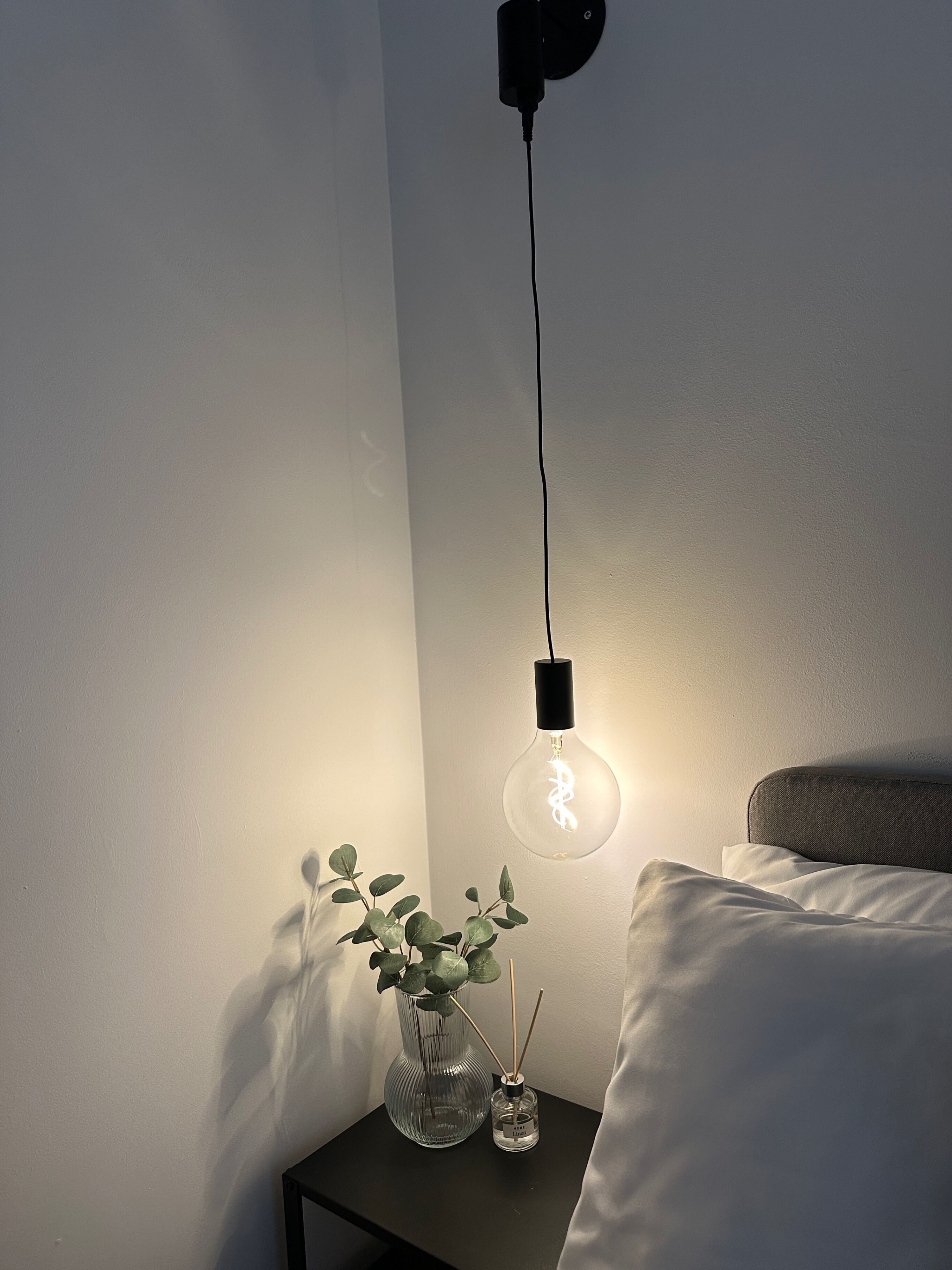 Une table de chevet sur laquelle se trouve un vase en verre transparent contenant des feuilles vertes et des diffuseurs de parfum à bâtonnets, une lampe noire avec un abat-jour en verre transparent, et un oreiller blanc sur un lit.