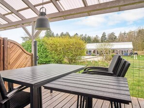 Un espace extérieur abrité, équipé d’une table et de chaises, d’un luminaire suspendu, et offrant une vue sur un bâtiment en arrière-plan.