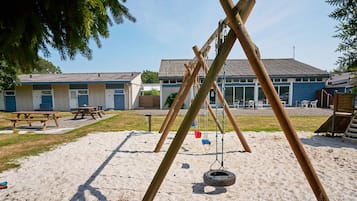 Un parque infantil con columpios, un neumático colgante, mesas de picnic y un edificio con tejado de azulejos al fondo.